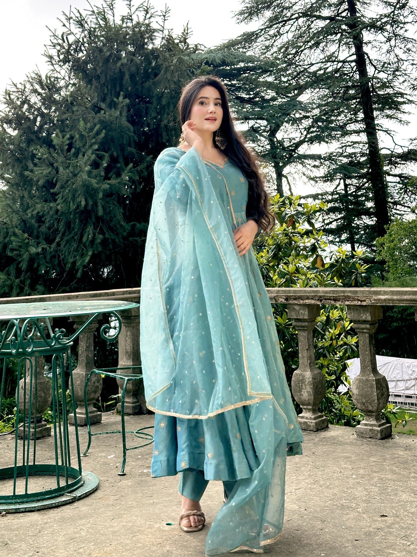 Woman in a light blue traditional outfit standing outdoors with greenery in the background