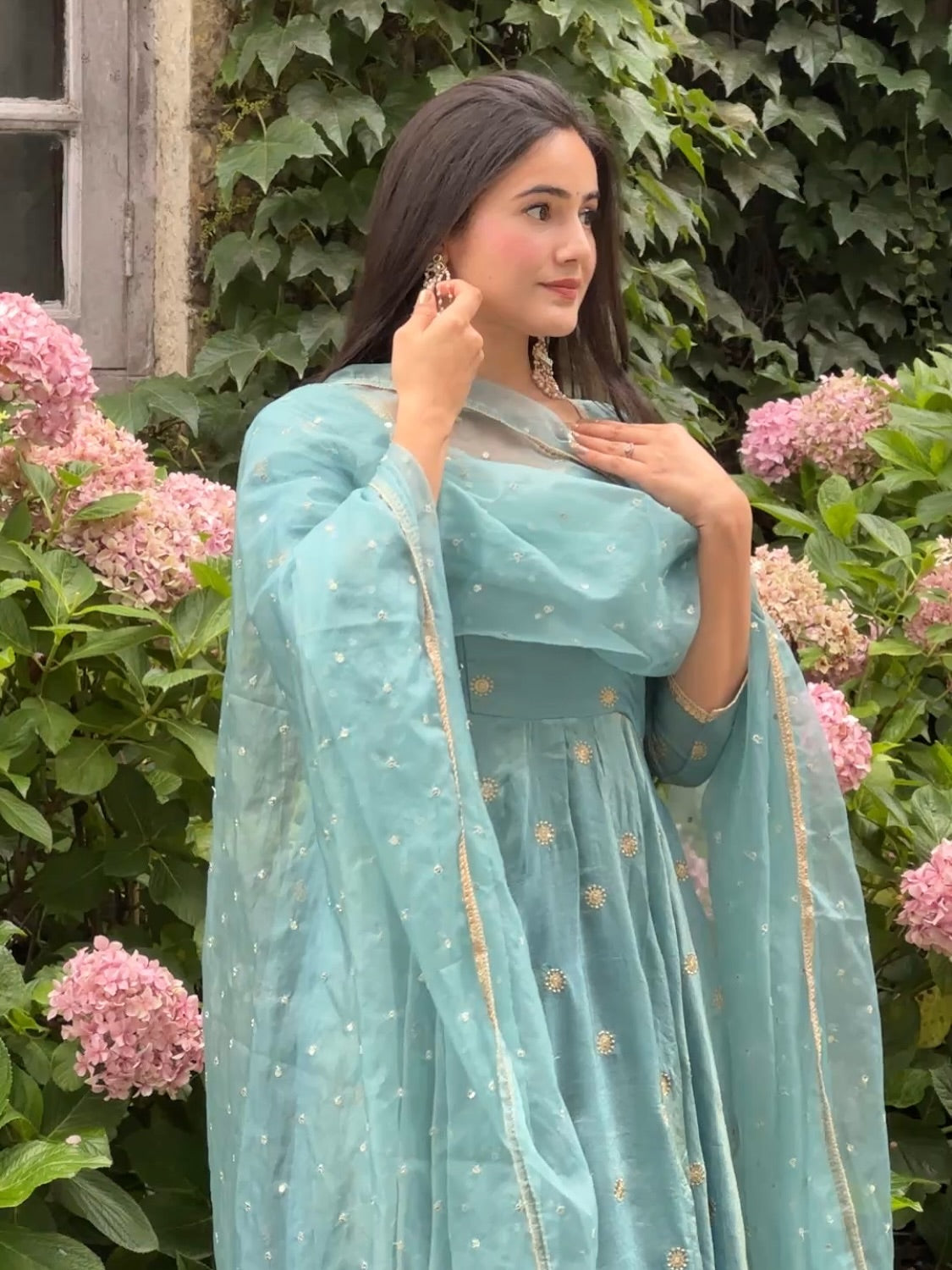 Woman in a light blue traditional outfit standing in front of pink flowers and green foliage