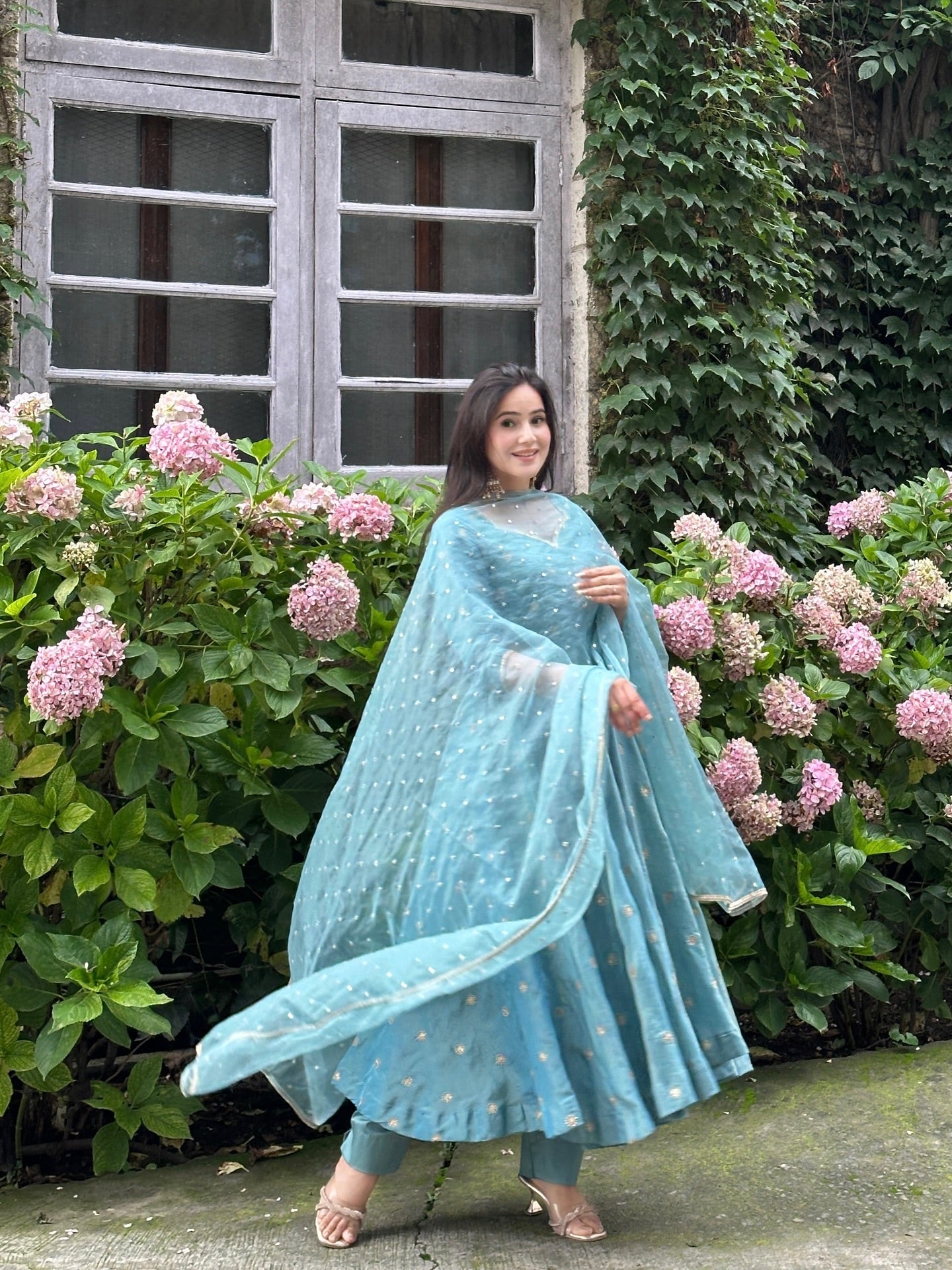 Woman in a light blue traditional outfit standing in front of pink flowers and a building.
