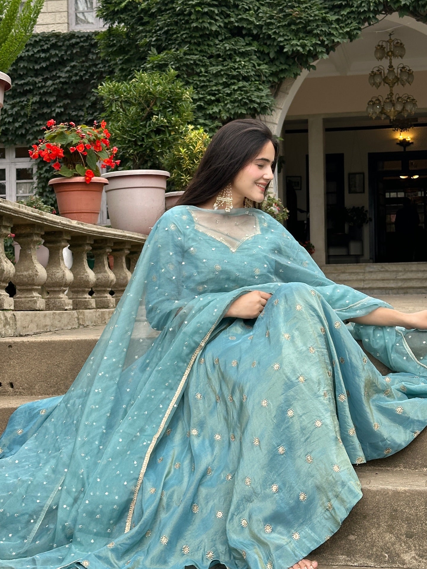 Woman in a light blue saree sitting outdoors with plants and a building in the background