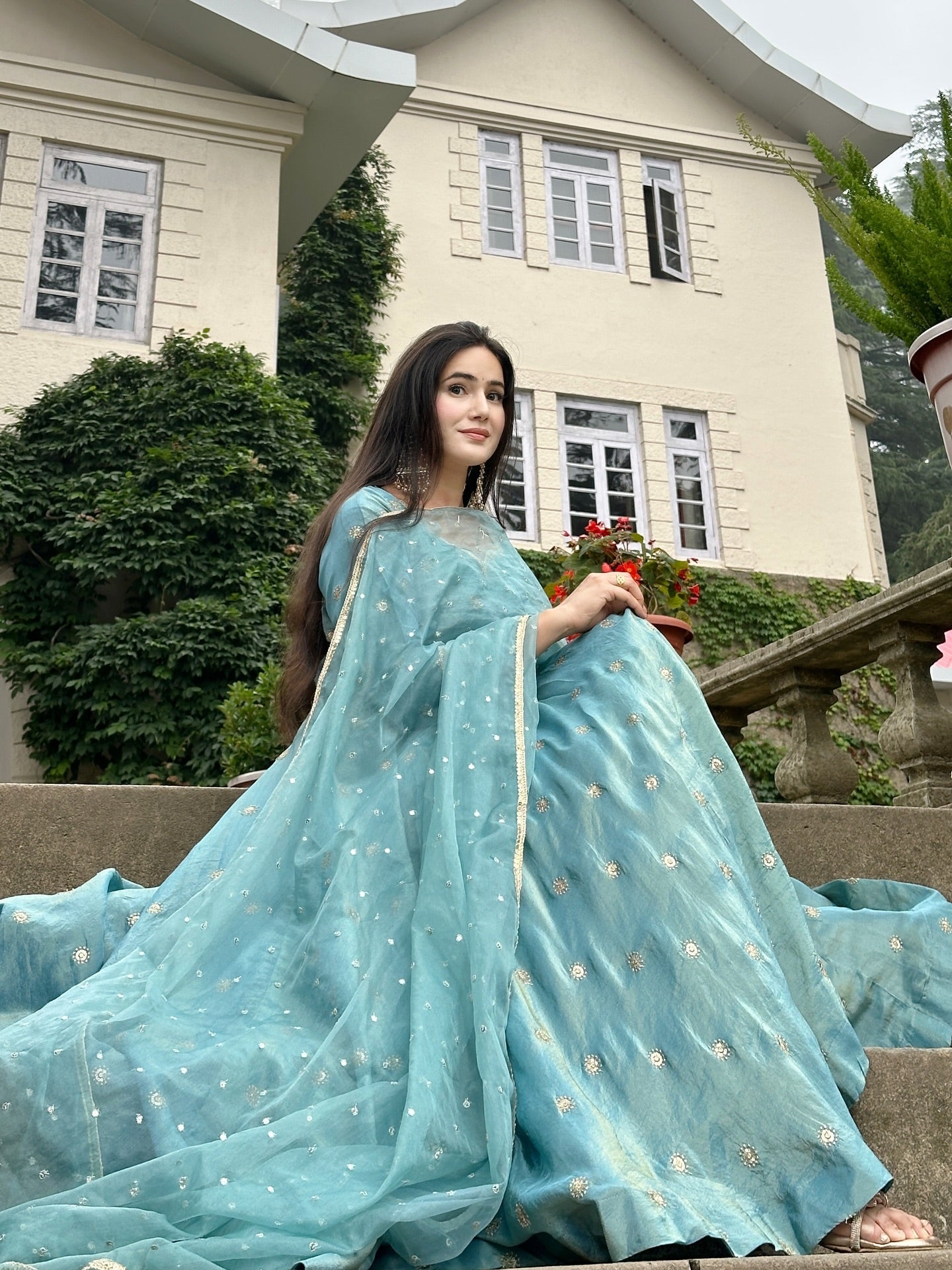 Woman in a light blue saree sitting on steps with a building and greenery in the background