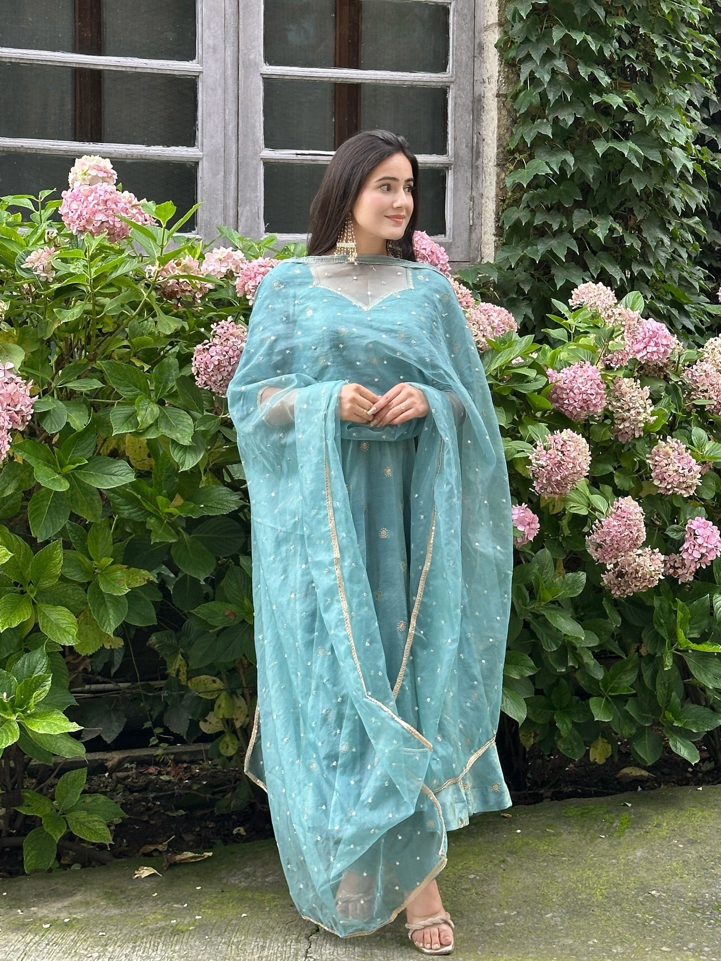 Woman in a light blue saree standing in front of a building with greenery and flowers.