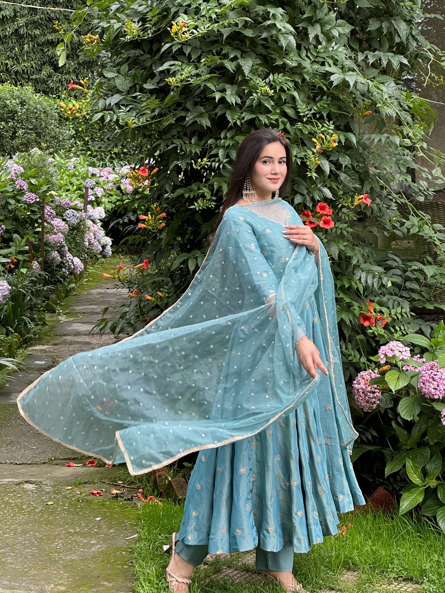 Woman in a light blue traditional outfit standing in a garden with greenery and flowers.