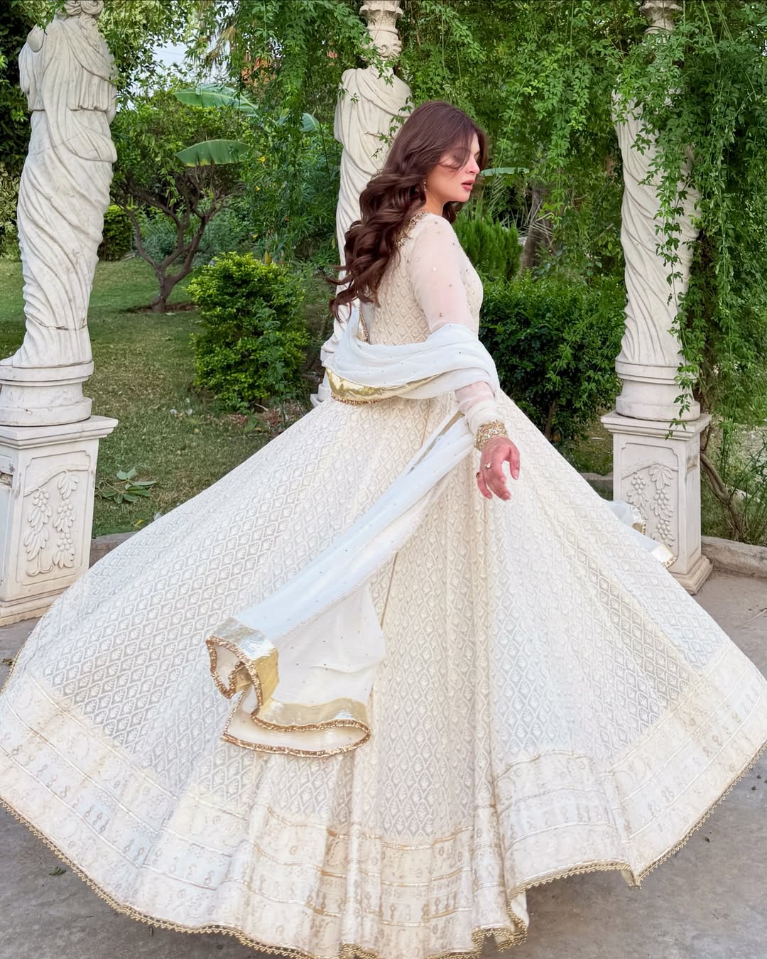 Woman in a white traditional outfit with gold details standing in a garden with statues.