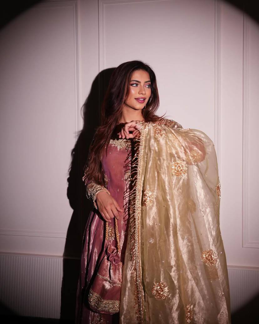 Woman wearing a traditional outfit with a gold dupatta against a white wall.