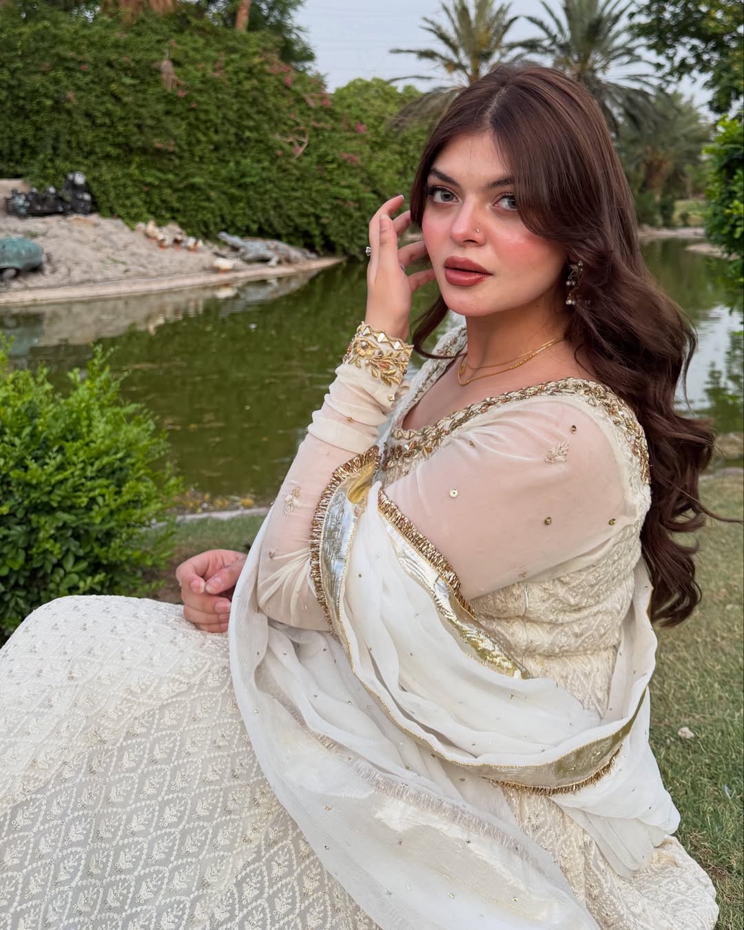 Woman in a white and gold traditional outfit sitting by a pond with greenery in the background