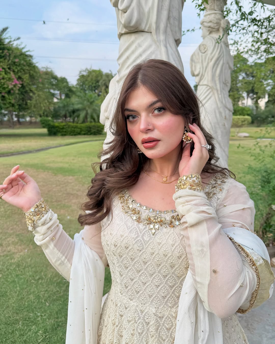 Woman in a white and gold traditional outfit standing outdoors with greenery and statues in the background