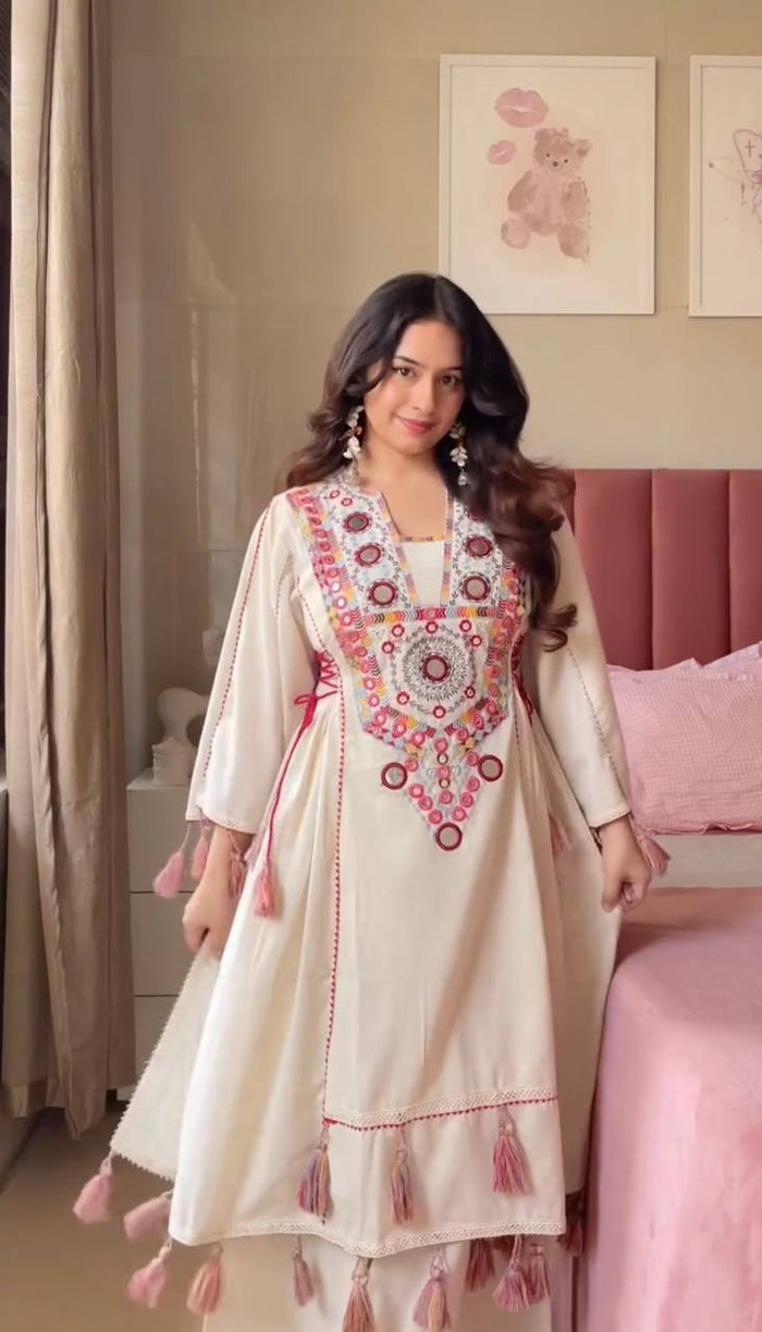 Woman in a cream-colored traditional outfit with floral embroidery in a bedroom setting.