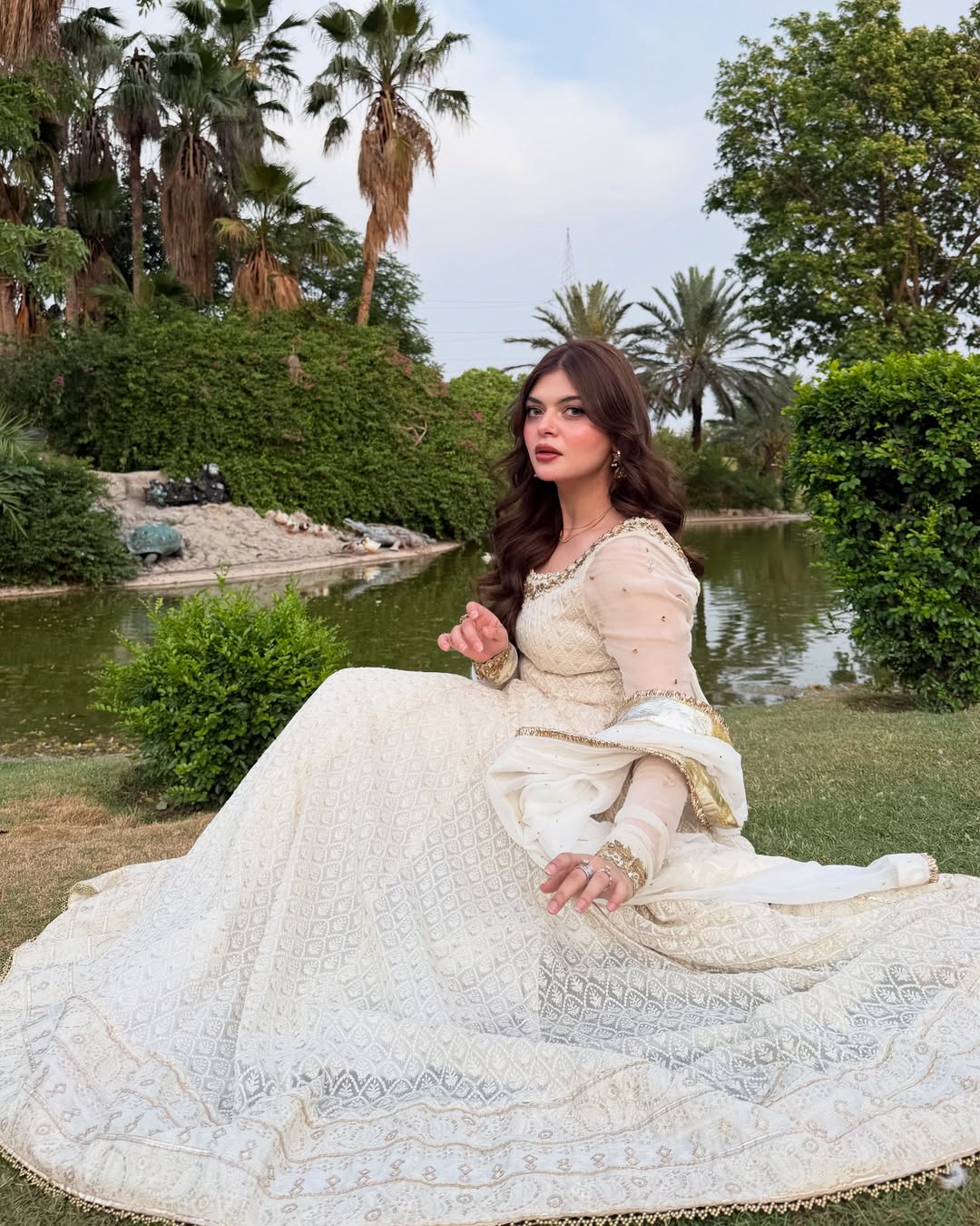 Woman in a white traditional outfit sitting outdoors with greenery and water in the background