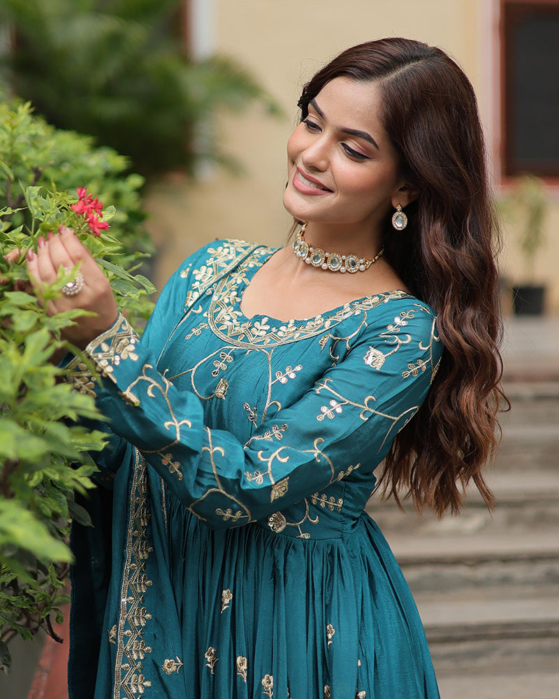 Woman in a teal embroidered dress standing outdoors with greenery.