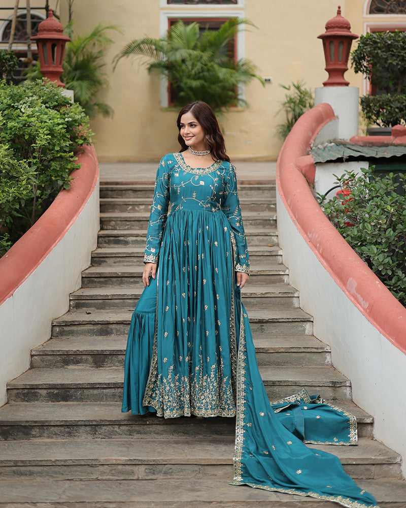 Woman in a teal embroidered dress standing on outdoor steps with greenery and a building in the background