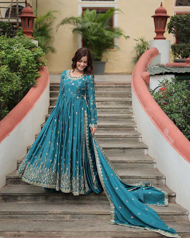 Woman in a teal embroidered dress standing on outdoor steps with greenery and a building in the background