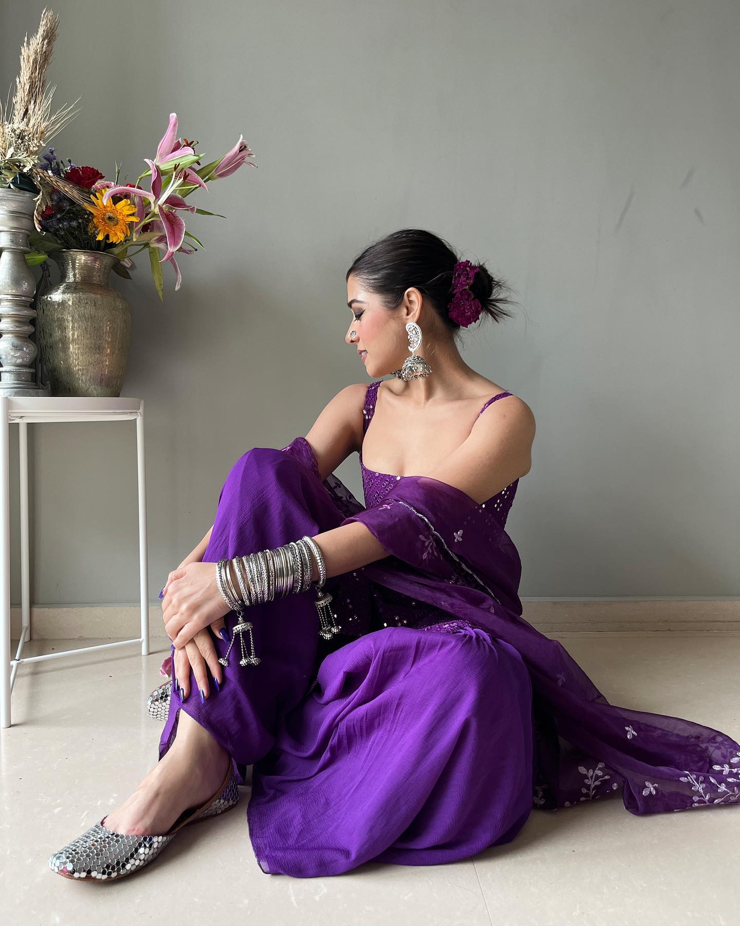 Woman in a purple saree sitting on the floor with a vase of flowers in the background