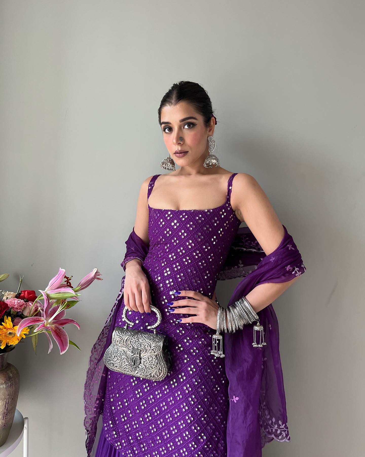 Woman in a purple traditional outfit holding a silver purse against a plain background