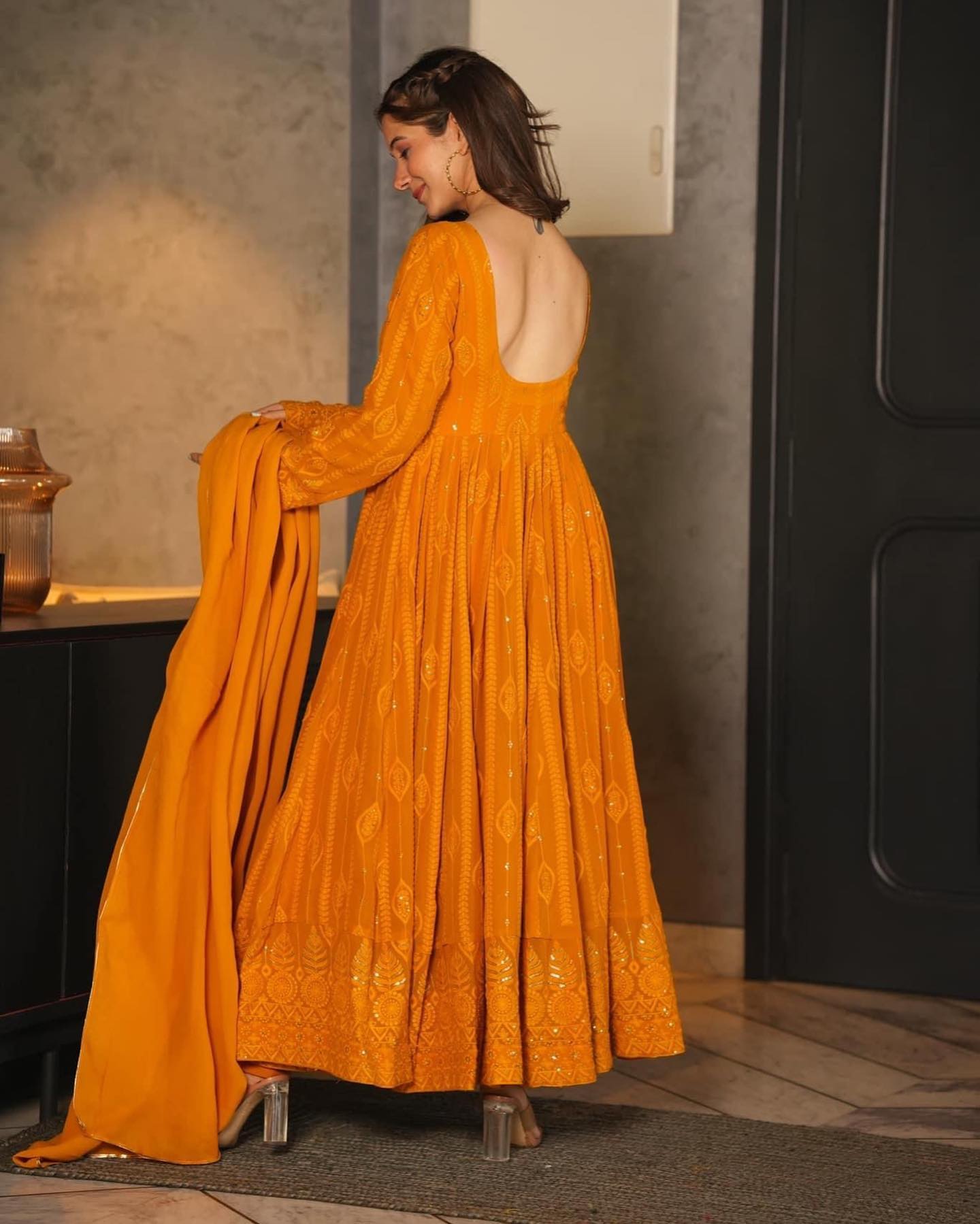 Woman in a long, mustard yellow dress with a backless design, standing indoors.