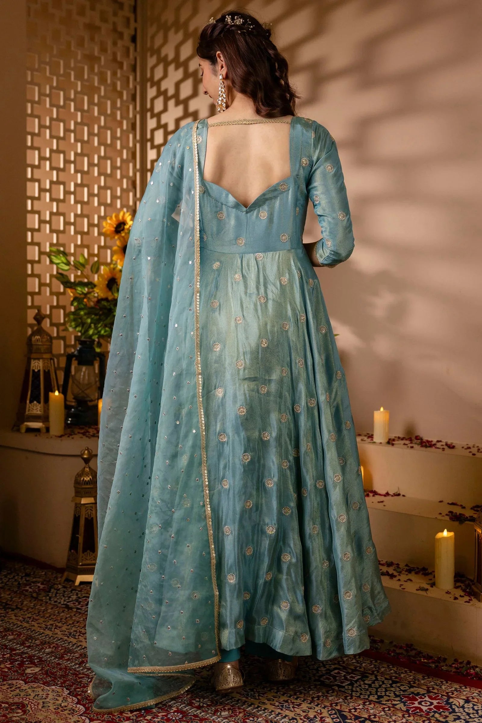 Woman in a light blue traditional outfit with a floral pattern, standing in a decorated room.