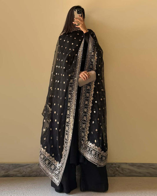 Person wearing a black traditional outfit with a patterned scarf against a beige wall.