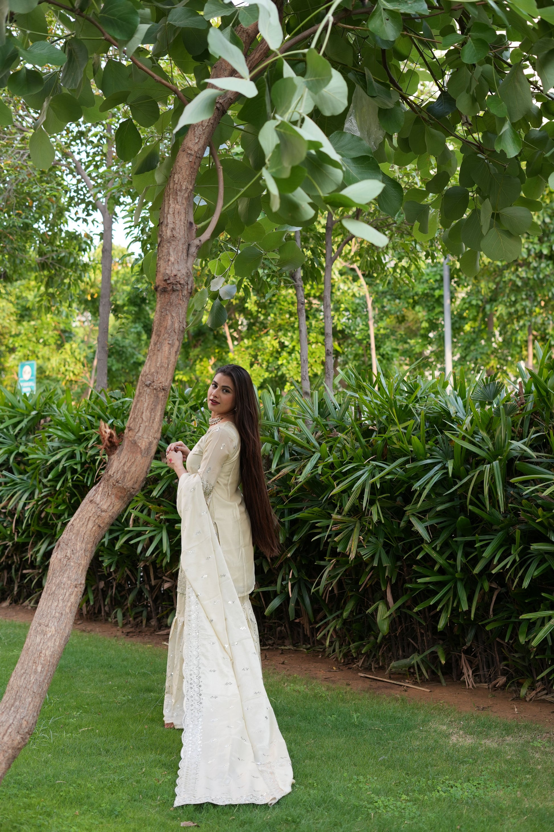 Woman in a white saree standing under a tree in a garden