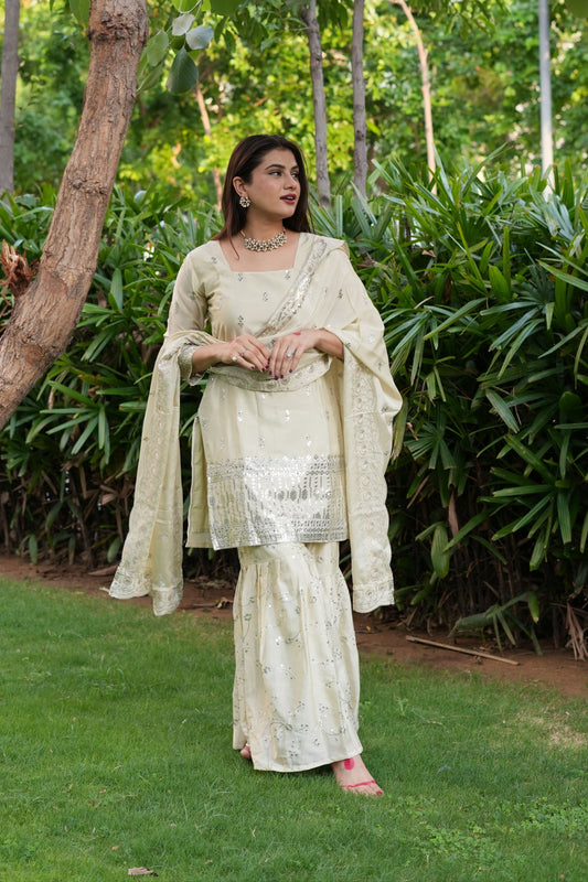 Woman in a light-colored traditional outfit standing in a natural setting with greenery.