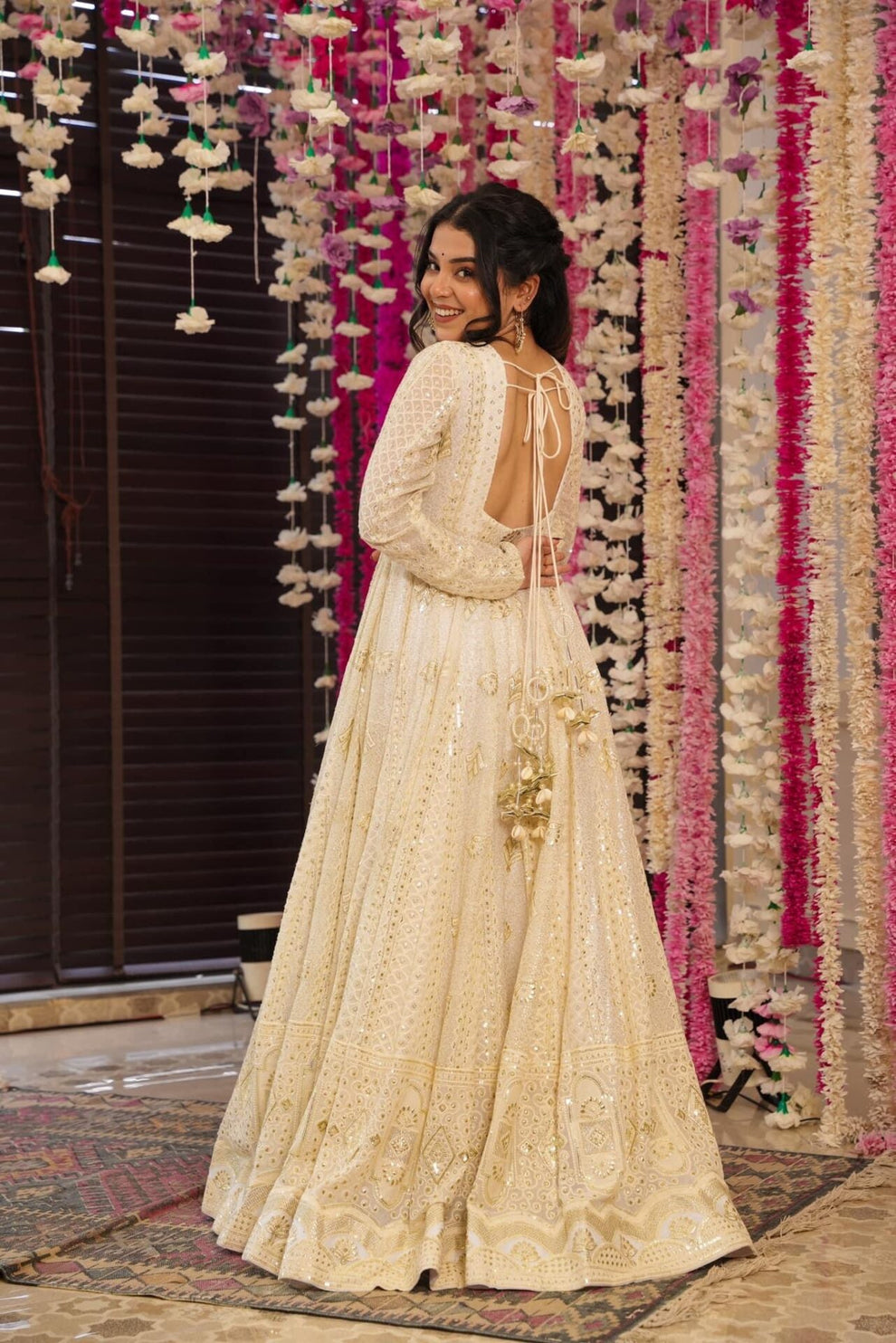 Woman in a cream embroidered dress standing in front of a decorative floral wall.
