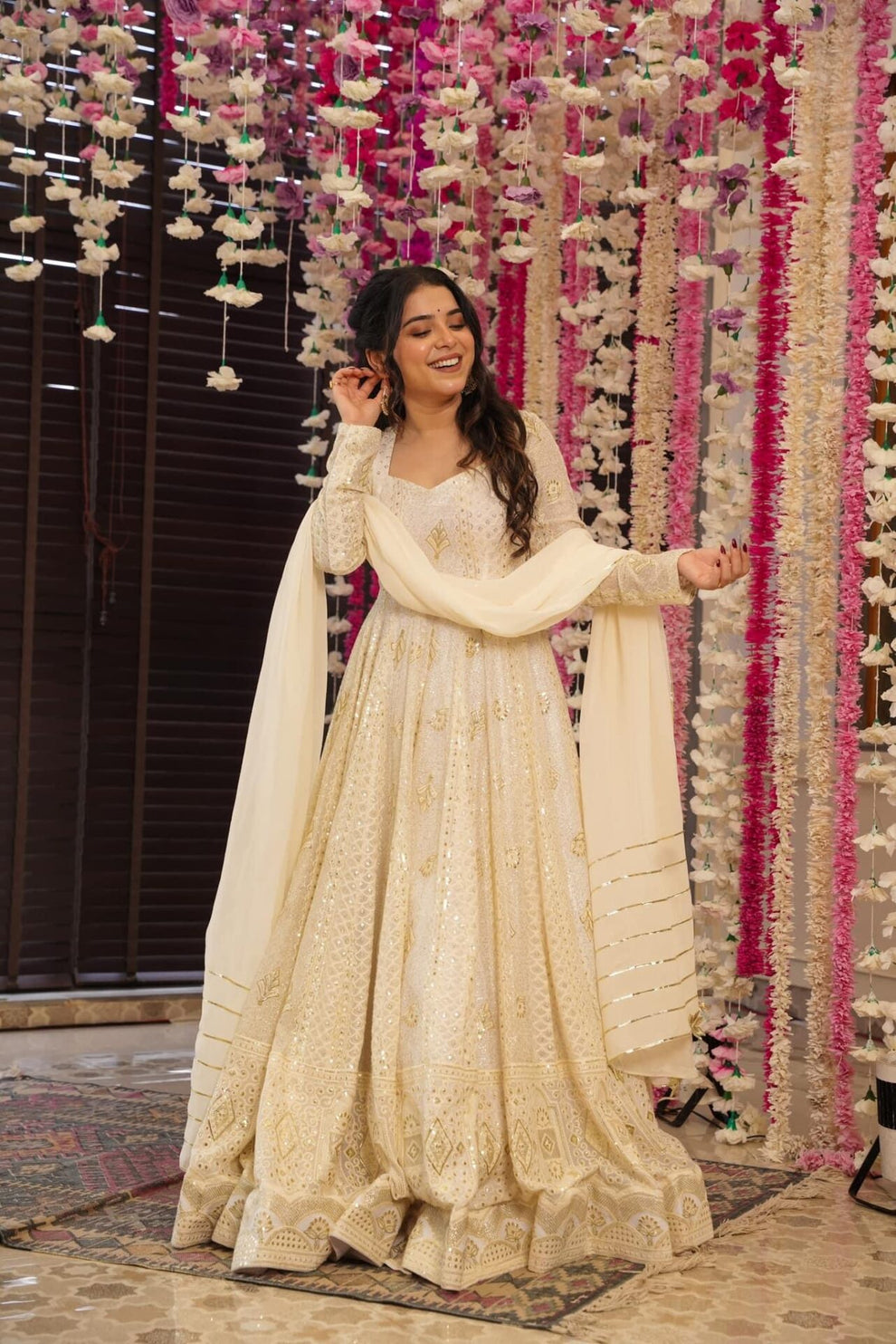 Woman in a cream traditional outfit standing in front of a decorative floral wall.