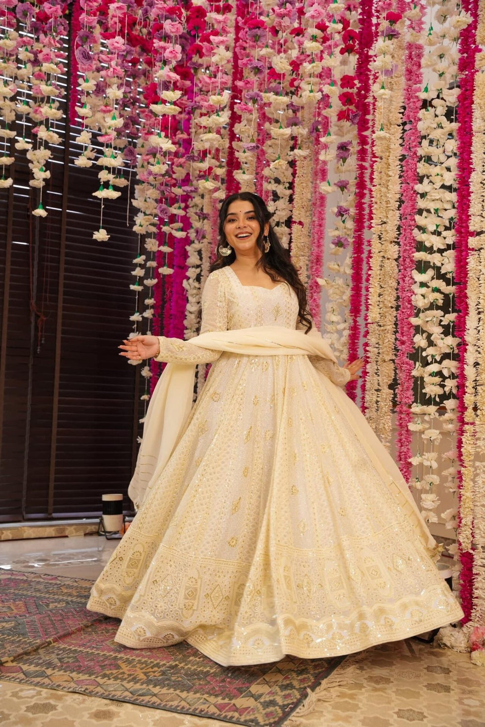 Woman in a cream traditional outfit standing in front of a decorative floral wall.