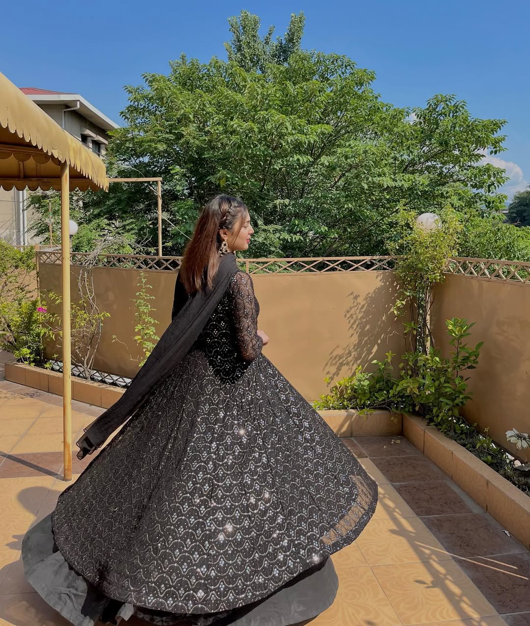 Woman in a black traditional outfit standing outdoors with greenery in the background