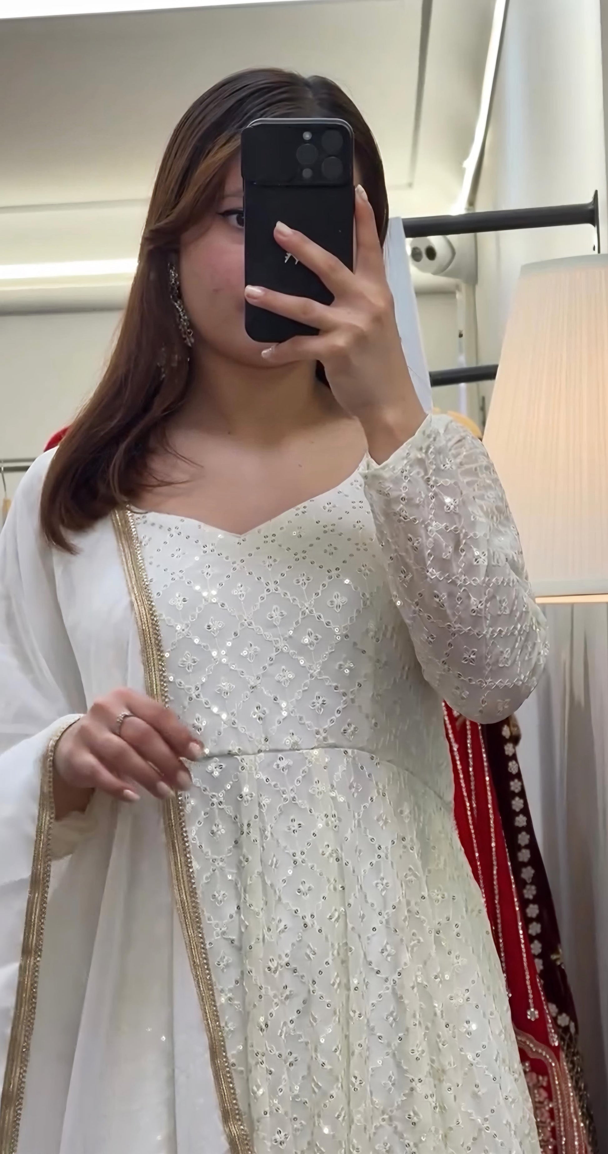 Person wearing a white traditional outfit with gold details, taking a mirror selfie.