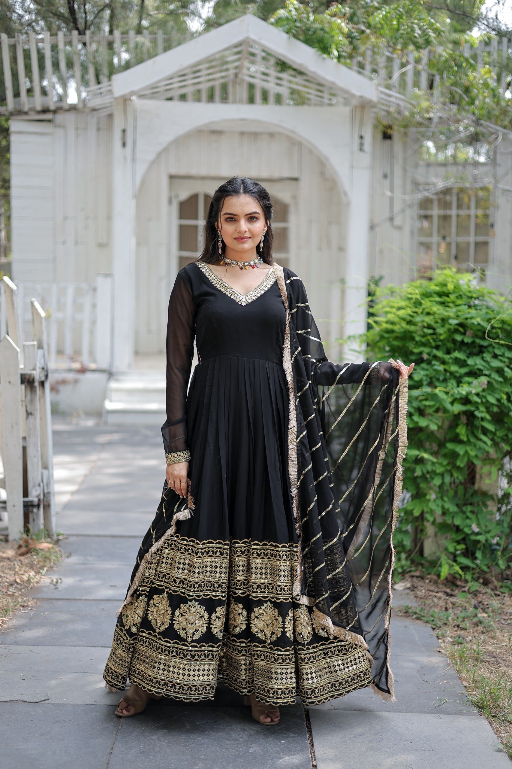 Woman in a black and gold traditional outfit standing outdoors with a white structure and greenery in the background.
