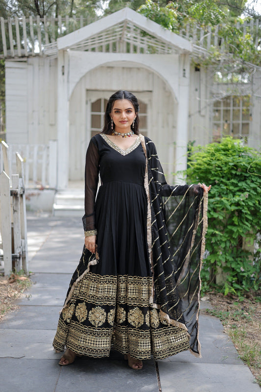 Woman in a black and gold traditional outfit standing outdoors with a white structure and greenery in the background.