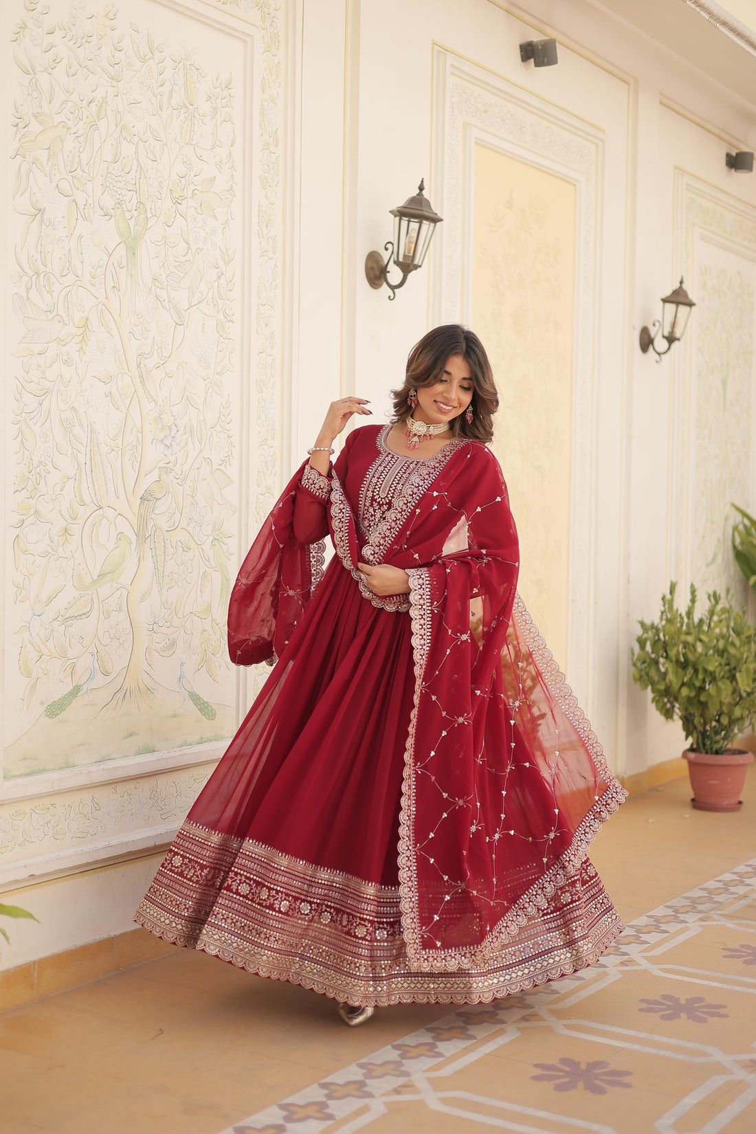 Woman in a red traditional outfit standing in a decorated room with white walls and a plant.