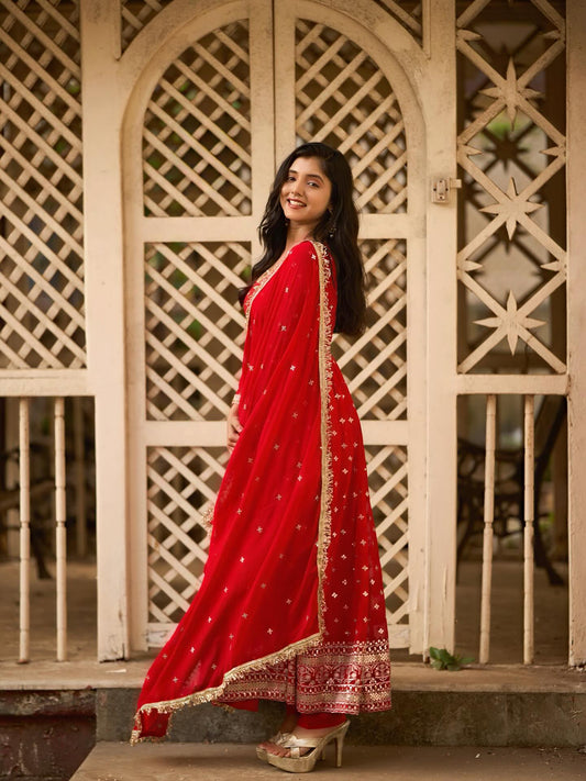 Woman in a red saree with white patterns standing in front of a decorative wooden gate.