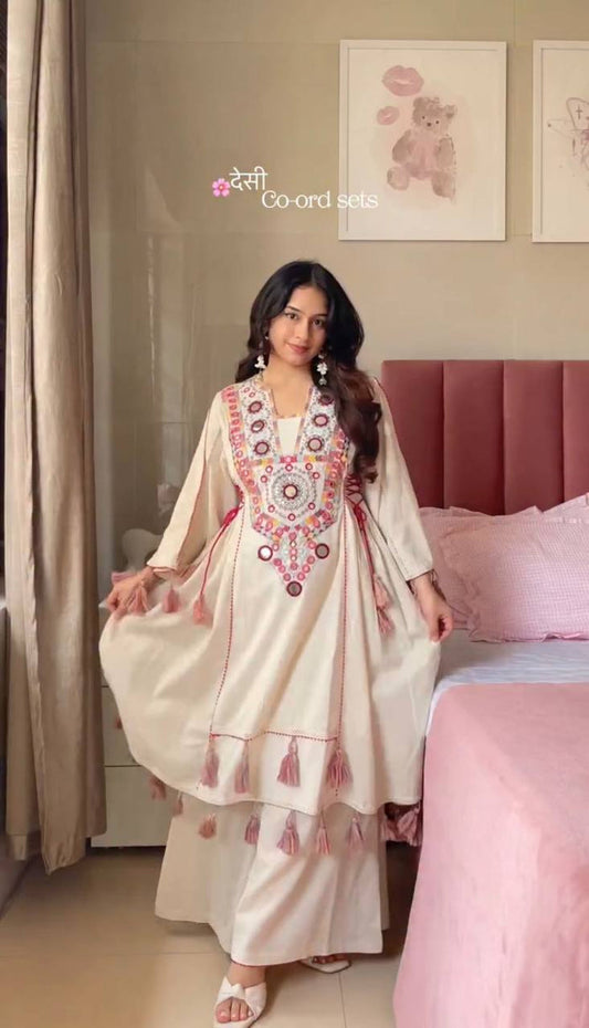 Woman in a cream-colored embroidered outfit standing in a bedroom with pink bedding.